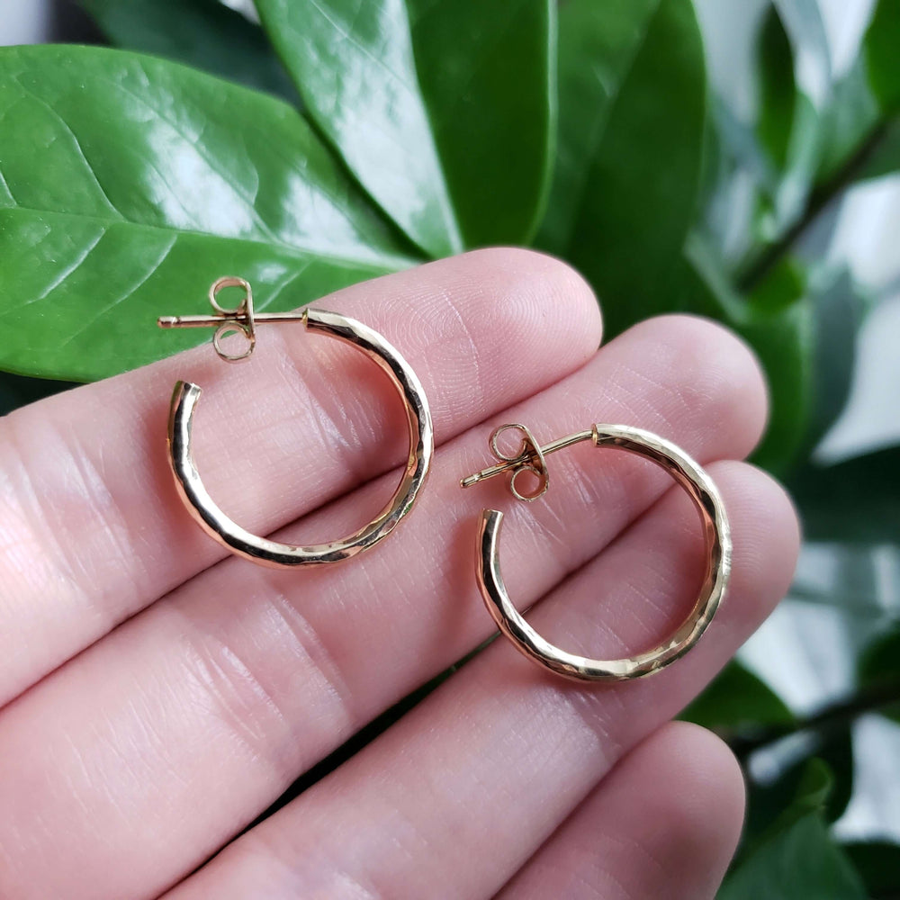 Hand holding hammered 14K yellow gold hoop earrings with green leaves in background