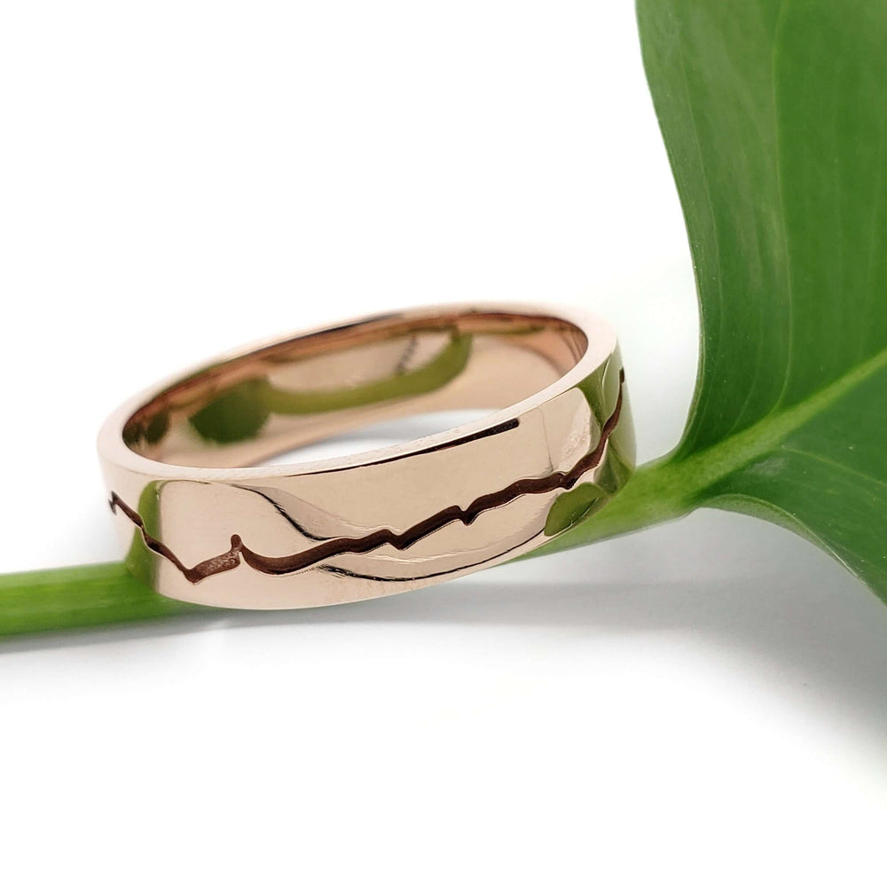 14k rose gold mountain range wedding ring resting on a green leaf, polished finish