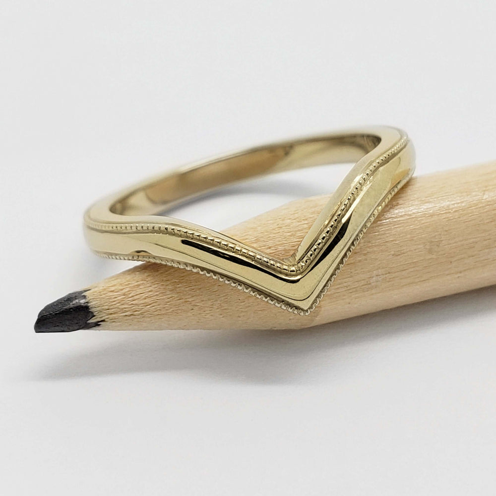 14kt yellow gold chevron wedding ring with millgrain resting on a pencil