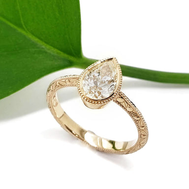 A gold ring with an engraved band and a pear-cut reclaimed diamond, styled on a white background with a green leaf prop.