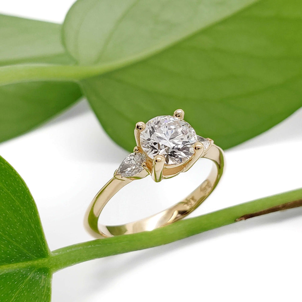 14kt yellow gold engagement ring with round lab diamond and pear side stones, displayed on green leaves