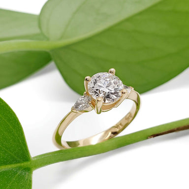 14kt yellow gold engagement ring with round lab diamond and pear side stones, displayed on green leaves