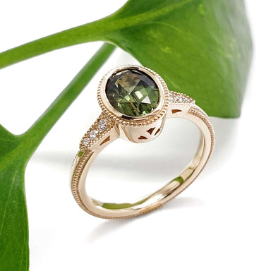 Handcrafted 14kt yellow gold ring with green oval sapphire and diamond accents, beside a green leaf