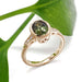 Handcrafted 14kt yellow gold ring with green oval sapphire and diamond accents, beside a green leaf