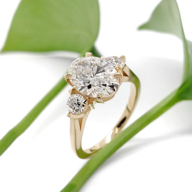 14kt yellow gold engagement ring with oval lab diamond and two round lab diamonds, shown with green leaves