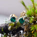 Pair of 14kt yellow gold lab emerald stud earrings resting on moss and lichen