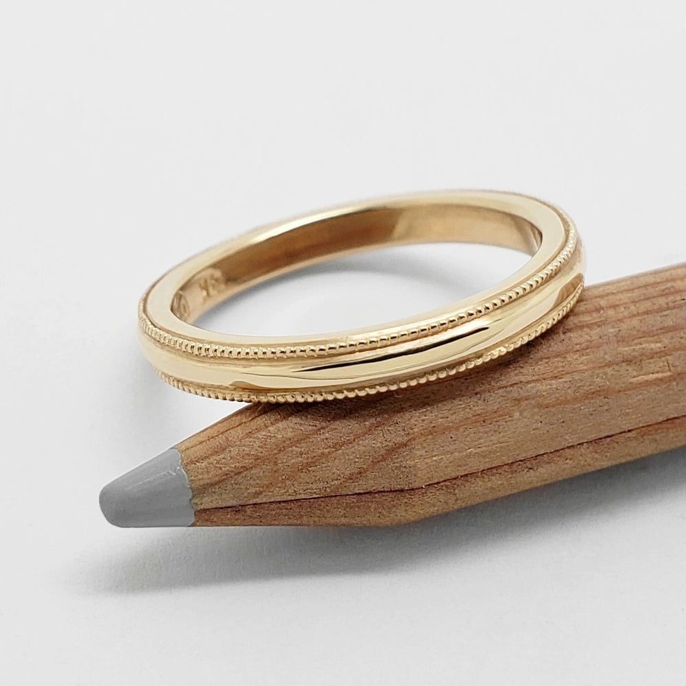 14kt yellow gold millgrain wedding ring resting on a wooden pencil