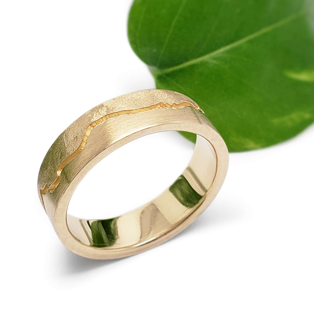14kt yellow gold mountain range wedding ring with brushed and burr textures beside a green leaf
