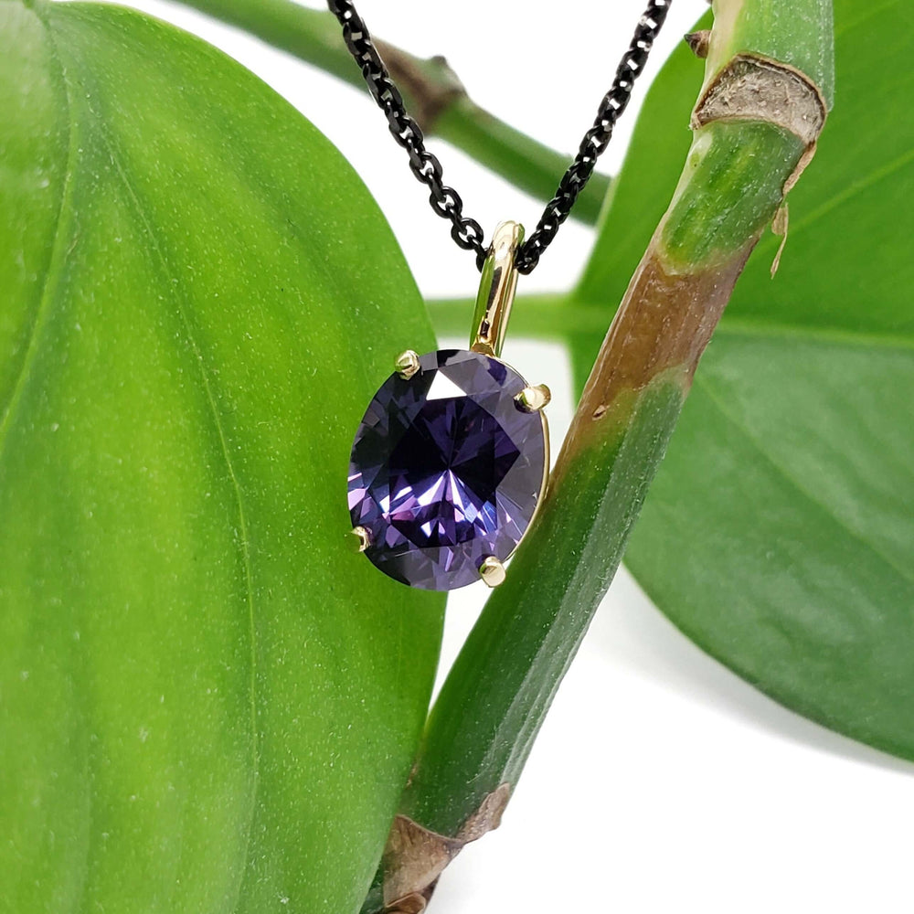 Oval lab grown purple sapphire pendant in 14kt yellow gold on a black chain, displayed on a green plant