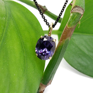 Oval lab grown purple sapphire pendant in 14kt yellow gold on a black chain, displayed on a green plant