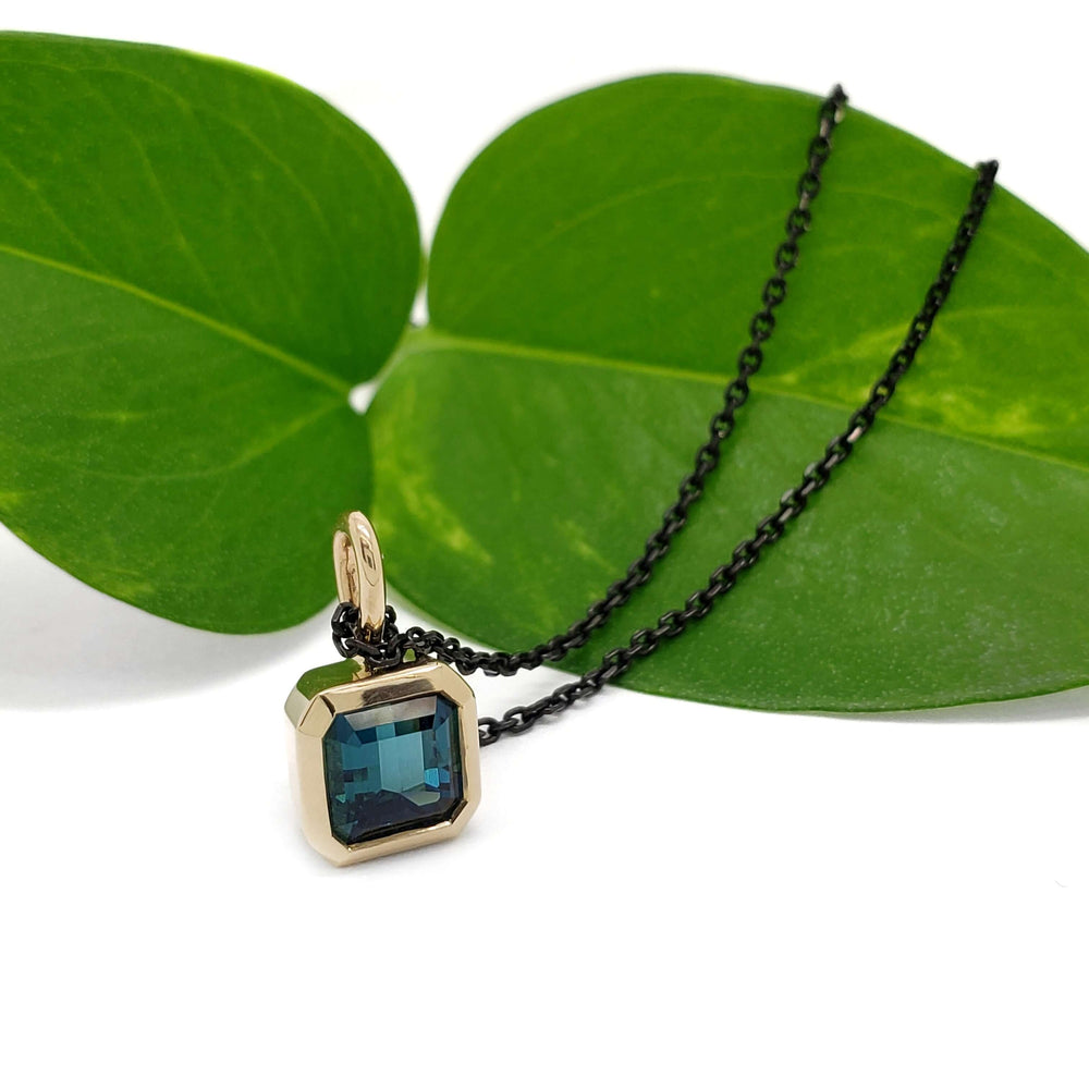 14kt yellow gold teal tourmaline bezel pendant on black chain, styled with green leaves
