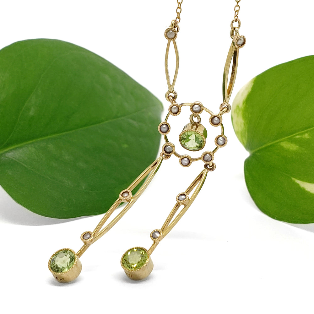 Gold-tone dangle necklace with green gemstone drops and small clear stones, styled with two green leaves on a white background.