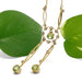 Gold-tone dangle necklace with green gemstone drops and small clear stones, styled with two green leaves on a white background.