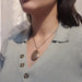 A close-up of a woman wearing a gold-toned oval pendant necklace with a textured, glittery surface, paired with a light sage green ribbed knit top.