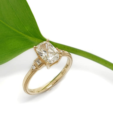 A yellow gold ring with a cushion-cut clear center stone and small side stones, styled on a white background with a green leaf prop.