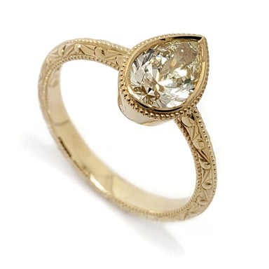 A gold ring featuring an oval-cut clear gemstone in a beaded bezel setting on an ornate, engraved band, shown on a white background.