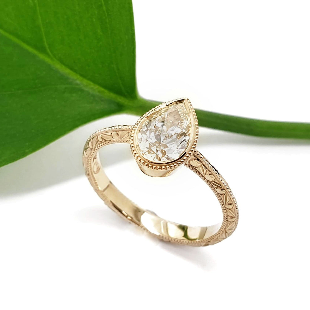 A gold ring with an engraved band and a pear-cut clear gemstone, styled on a white background with a green leaf prop.