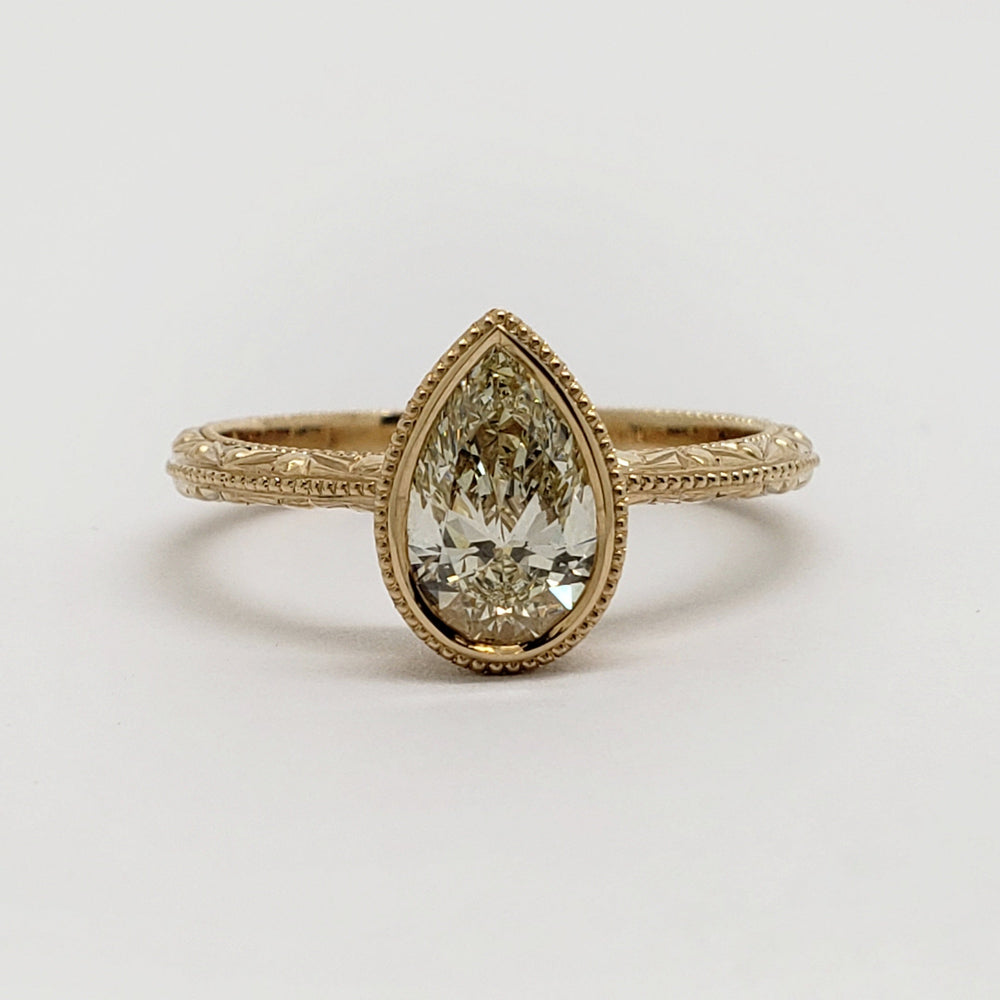 A yellow gold ring featuring a pear-cut diamond center stone in a bezel setting, with an ornate filigree band, shown on a plain light gray background.