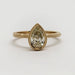 A yellow gold ring featuring a pear-cut diamond center stone in a bezel setting, with an ornate filigree band, shown on a plain light gray background.