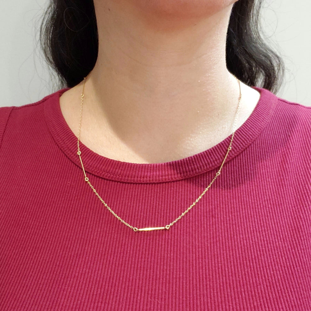 A delicate gold chain necklace with a small horizontal bar centerpiece, worn by a model wearing a magenta ribbed t-shirt.