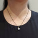A close-up of a person's neck and chest wearing a delicate dark chain necklace with a small gold bail and a clear faceted round pendant, paired with a black top.