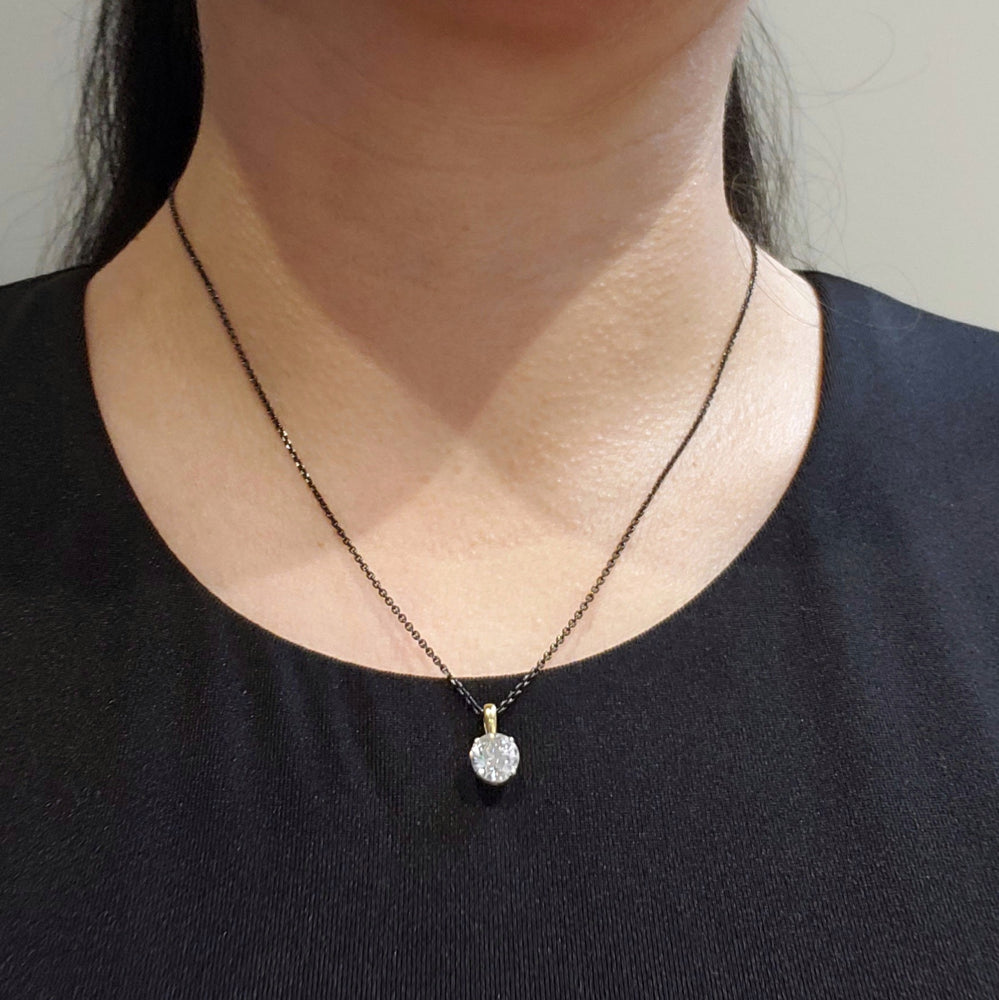 A close-up of a person's neck and chest wearing a delicate dark chain necklace with a small gold bail and a clear faceted round pendant, paired with a black top.