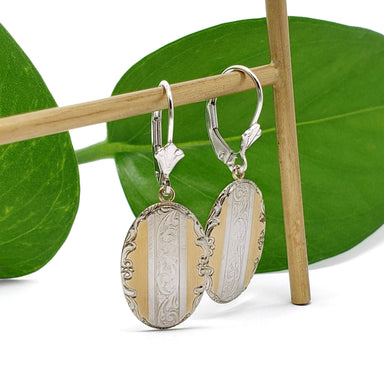 Antique oval earrings in yellow and white gold with engraved detail, hanging on a wooden rod