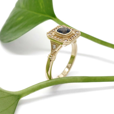 Art deco style yellow gold ring with blue Montana sapphire and diamond accents beside green leaves