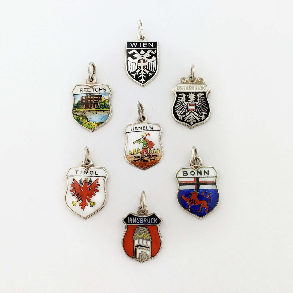 Seven assorted vintage silver crest pendants with city and region designs, displayed on a white background