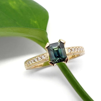 Emerald cut teal Australian sapphire and diamond engagement ring in yellow gold on white background with green leaf
