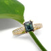 Emerald cut teal Australian sapphire and diamond engagement ring in yellow gold on white background with green leaf