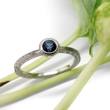 Bezel-set Montana sapphire ring in white gold with engraved band, shown with greenery