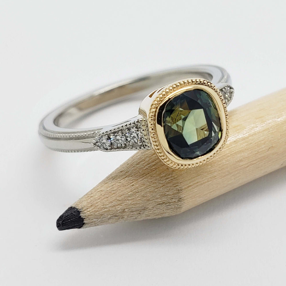 Bezel-set green sapphire and diamond engagement ring in 14k yellow and white gold, beside a pencil
