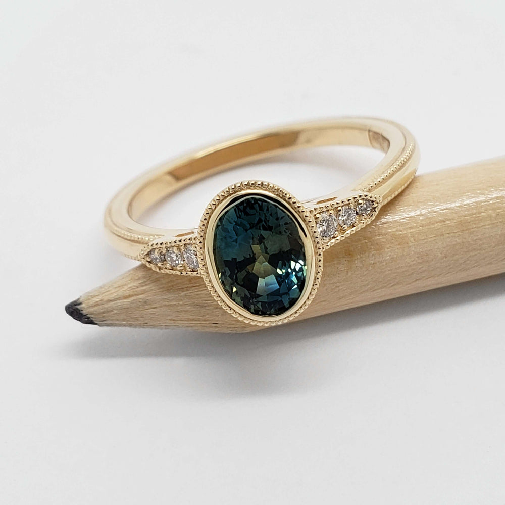 Bezel-set oval teal sapphire ring with diamond shoulders in yellow gold, displayed on pencil