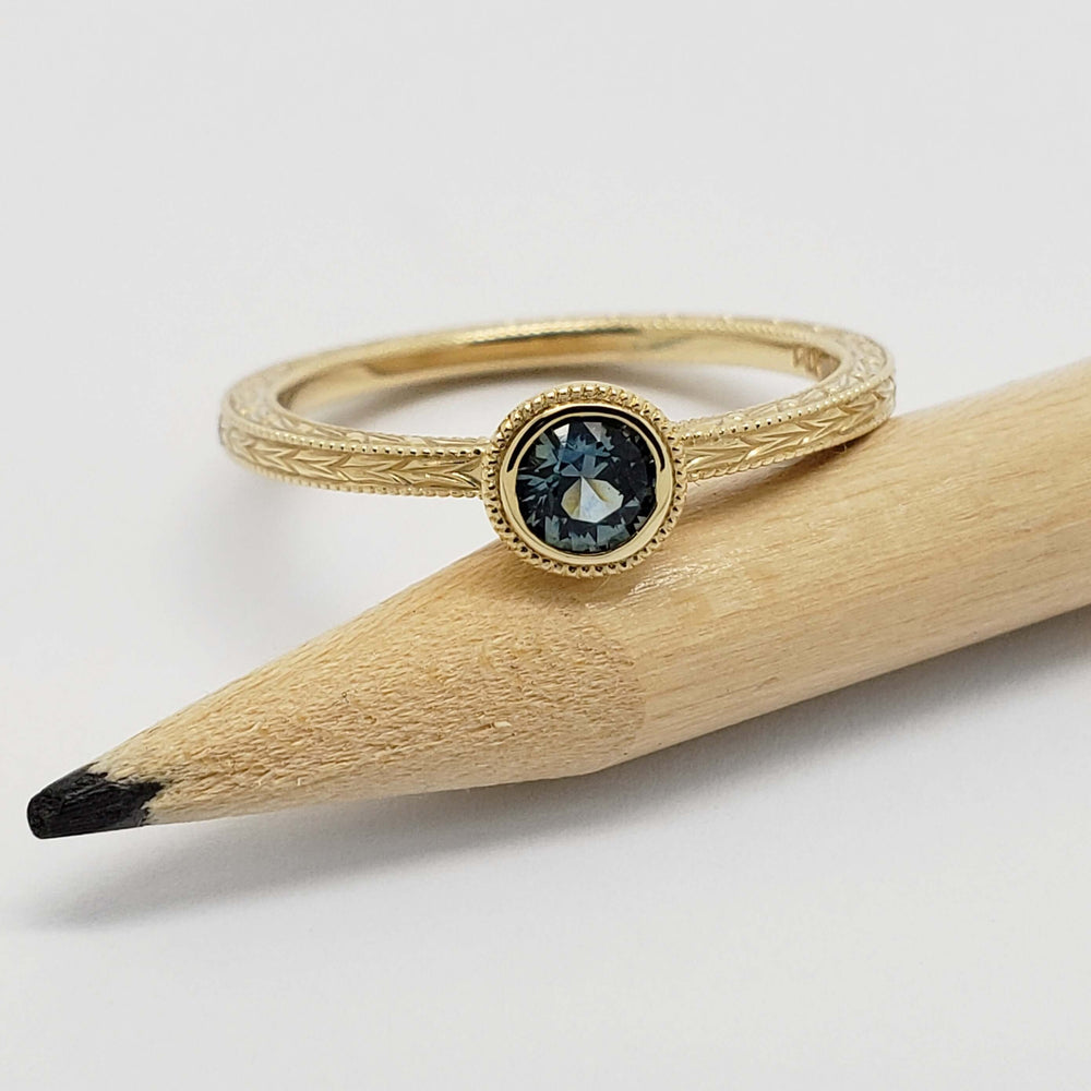 Bezel-set teal Montana sapphire ring in engraved yellow gold, displayed on a pencil
