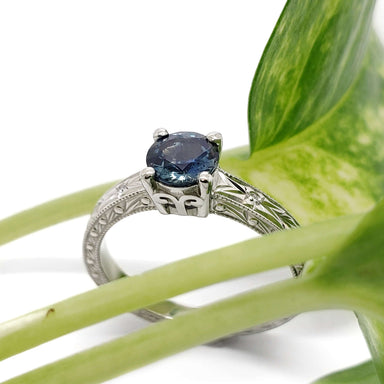 Blue Montana sapphire engagement ring in white gold with engraved details, beside green leaves