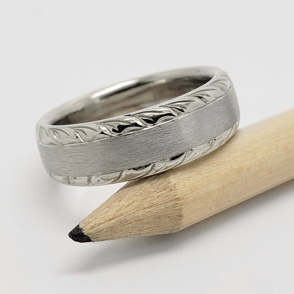 Brushed 14kt white gold ring with engraved wheat pattern edges resting on a pencil