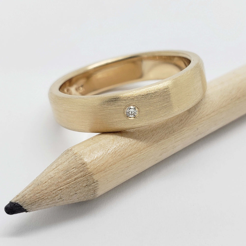 Brushed yellow gold wedding ring with flush-set Canadian diamond, shown on a pencil