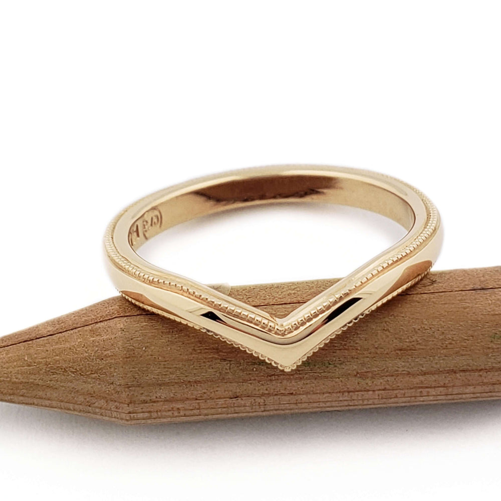 Chevron-shaped 14kt yellow gold wedding ring with millgrain, resting on wood