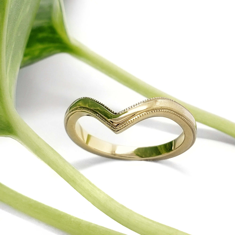 Chevron millgrain wedding ring in yellow gold displayed with green leaves