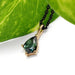 Close-up of a 14kt yellow gold pendant with shield-cut green sapphire on a black chain near a green leaf