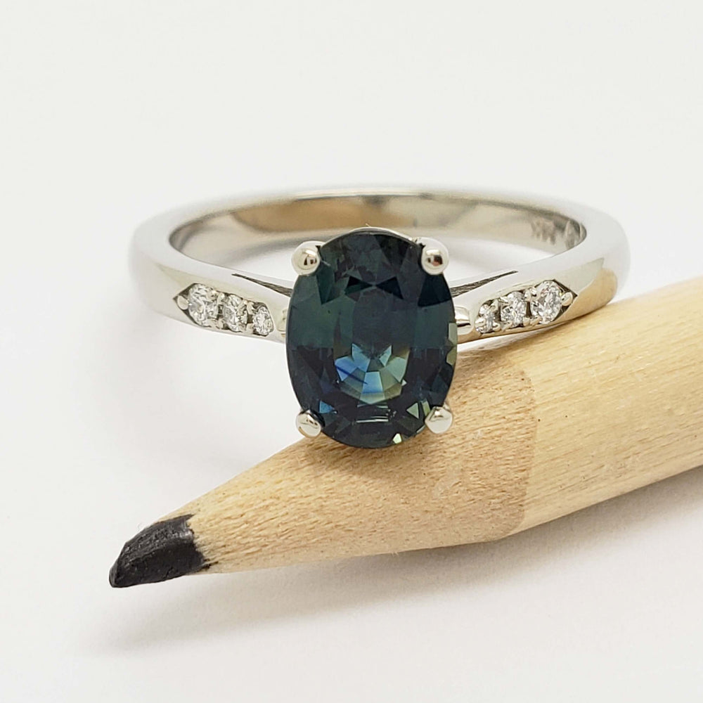 Close-up of oval blue-green sapphire ring with lab diamonds in white gold, beside pencil