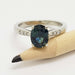Close-up of oval blue-green sapphire ring with lab diamonds in white gold, beside pencil