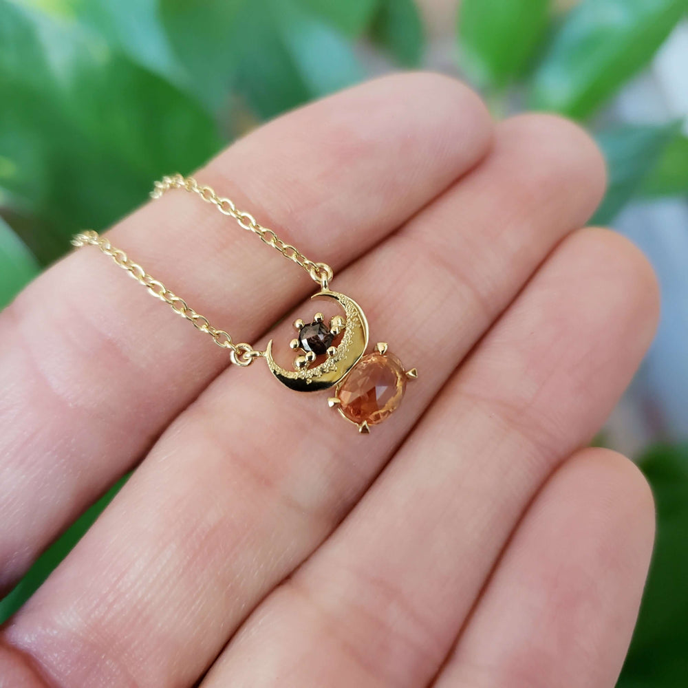 Hand holding 10kt yellow gold necklace with crescent moon, orange sapphire, and salt and pepper diamond