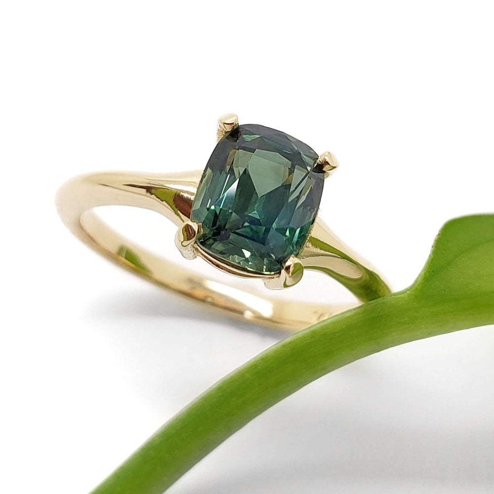 Cushion cut green sapphire engagement ring in yellow gold beside a green leaf