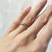 A close-up of a hand wearing a delicate gold ring with a small cluster of clear stones at the center, set against a softly blurred light background.