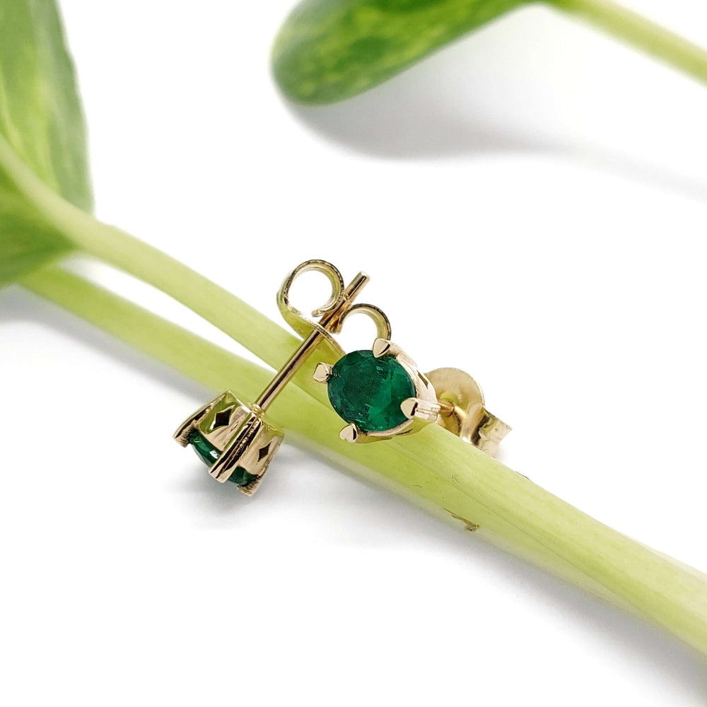 14kt yellow gold emerald stud earrings with heart prongs, shown resting on a green stem