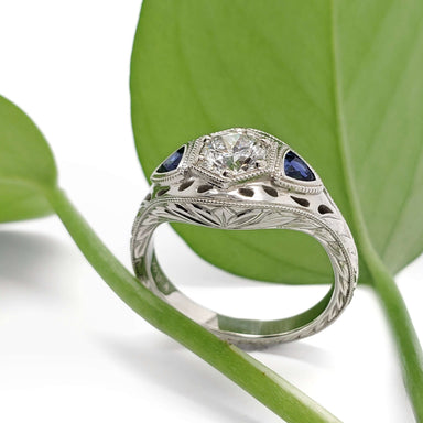 Hand-engraved white gold ring with lab diamond and blue sapphires, shown with green leaf background