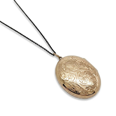 An oval gold locket pendant necklace with an engraved floral pattern, hanging from a thin black chain against a plain white background.