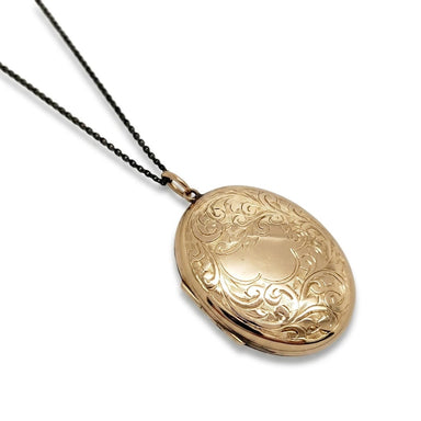 A gold oval locket necklace with intricate floral engravings, suspended from a thin black chain against a plain white background.